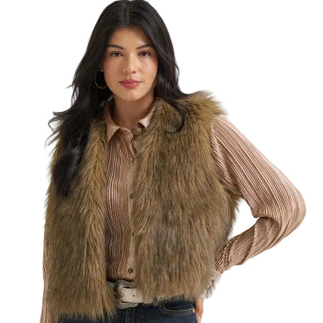 A woman modeling a brown faux fur vest over a beige pleated button-down shirt and dark blue jeans, photographed against a light gray studio background.